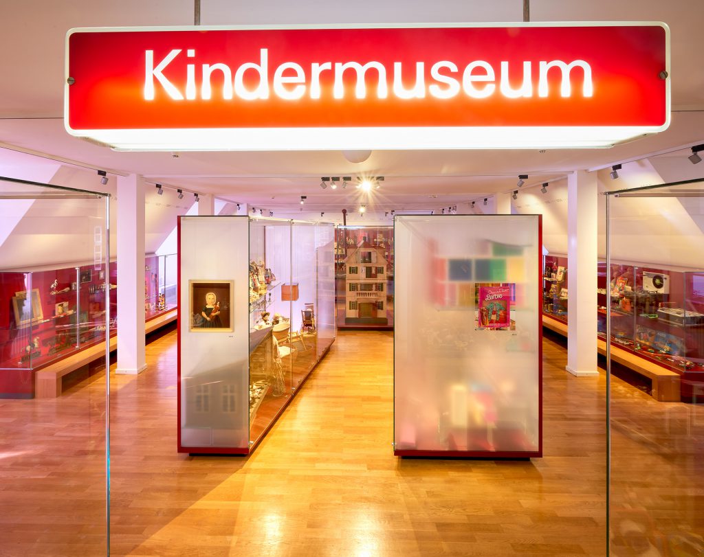 Blick auf Vitrinen mit Spielzeug. Oben hängt ein rotes beleuchtetes Schild: KIndermuseum.